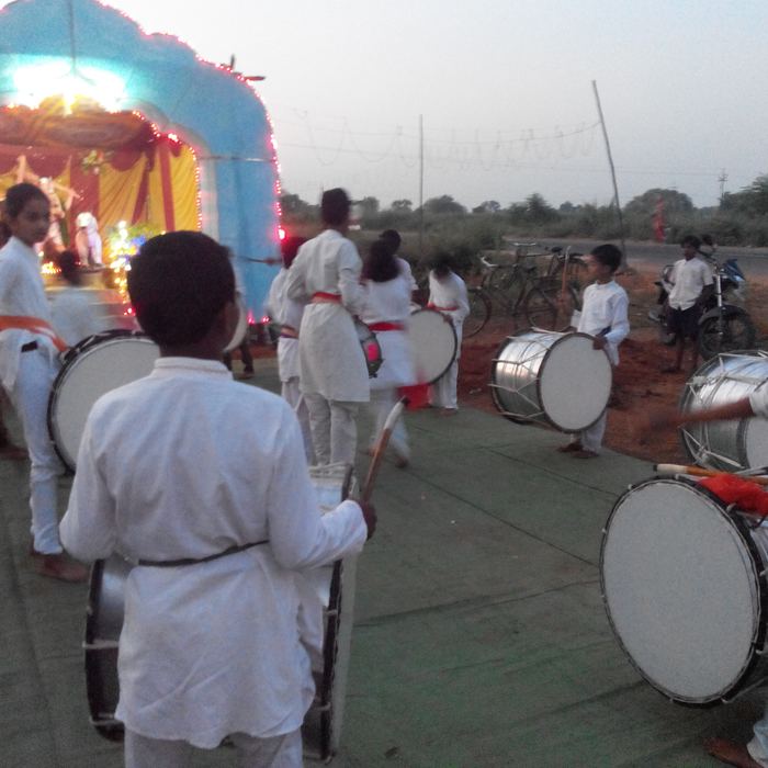 Students in white kurtas playing dhol drums in formation at dusk in front of an illuminated festival mandap