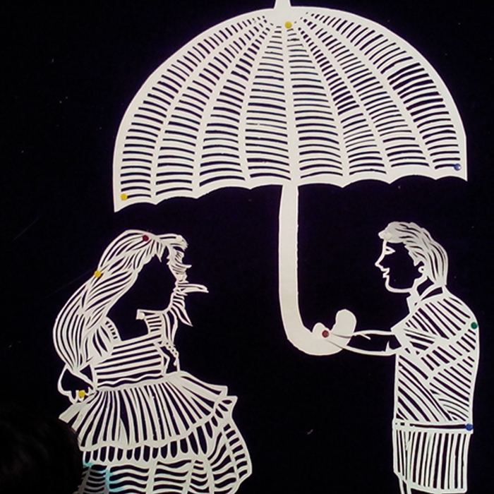 An intricate white paper cutting on a black background showing two figures under a large ribbed umbrella, cut from a single sheet