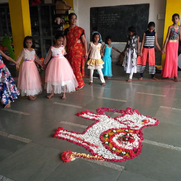 Girls in bright dresses holding hands in a circle around a flower rangoli while a teacher in a red saree leads the singing