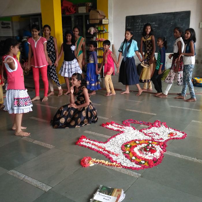 A girl kneeling beside a large peacock rangoli made of red and white flower petals, with classmates standing in a circle behind her