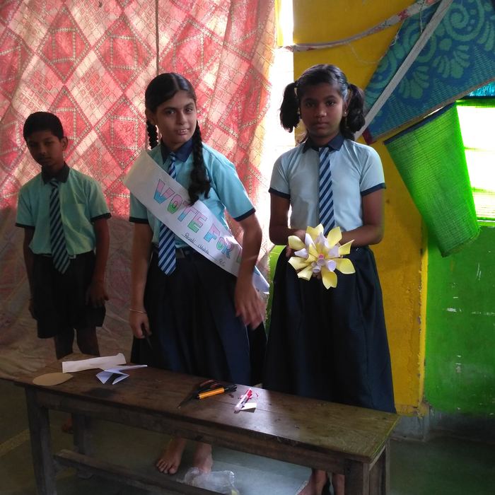 Two students standing at a wooden desk, one wearing a Vote For Me sash and the other holding up a yellow paper flower she has made