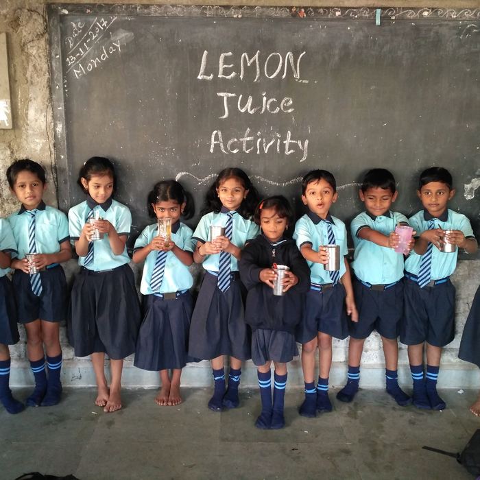 A line of young students in uniform holding steel tumblers of juice in front of a blackboard reading Lemon Juice Activity