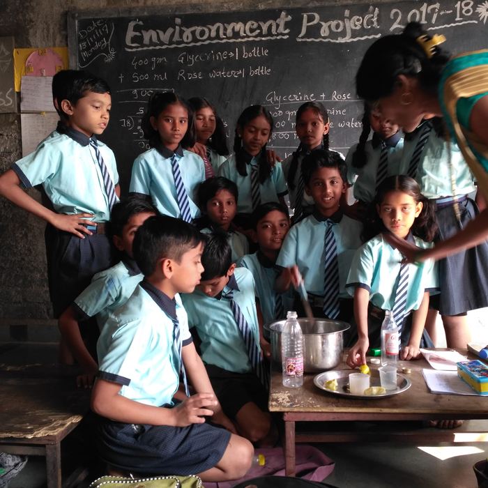 Students crowded around a steel mixing bowl and bottles of rose water and glycerine, with the recipe written on the blackboard behind them