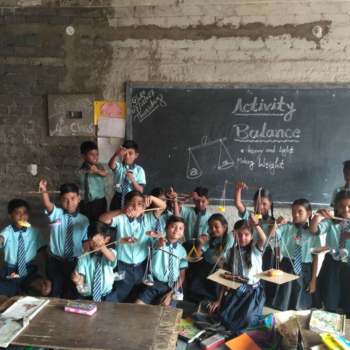 A whole class holding up balances they have built from sticks, string and small pans, in front of a blackboard reading Activity Balance