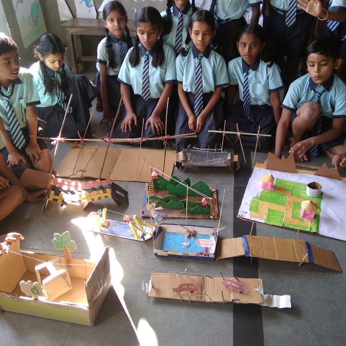Students sitting on the floor around a spread of handmade cardboard and skewer bridge models, including suspension and cable-stayed designs