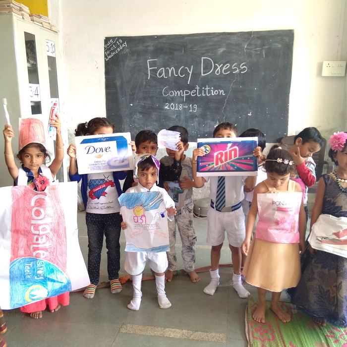 Small children in handmade paper costumes shaped like household product packets, standing before a blackboard reading Fancy Dress Competition