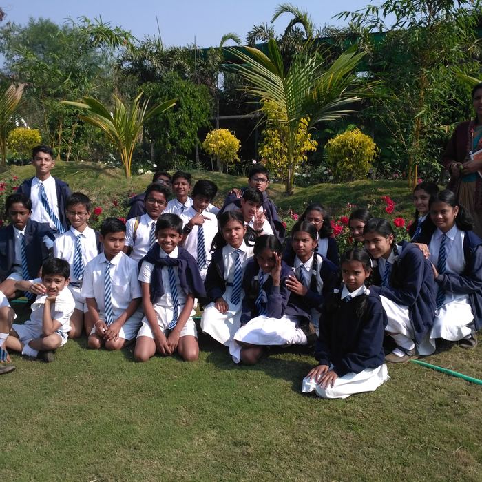 A class of students seated together on a lawn in front of rose beds and palms with their teacher standing at the side