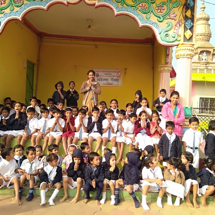 A large group of schoolchildren seated with palms joined in front of a decorated temple hall marked Yoga Kendra