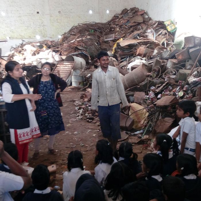 Students seated on the ground listening to a scrap dealer explain his work in front of large piles of sorted metal waste