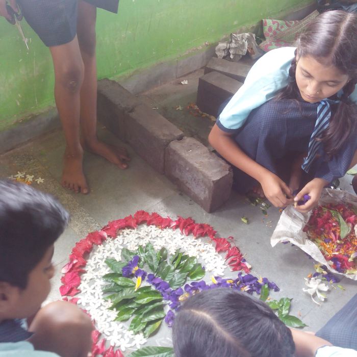 Children kneeling on a floor sorting flower petals from newspaper trays while filling in a circular rangoli of red, white and purple flowers