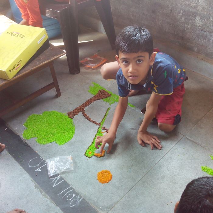 A boy kneeling on a classroom floor placing coloured grains into a chalked outline of a tree, with bags of pulses beside him