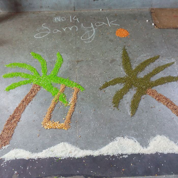 A finished grain rangoli of two coconut palms above a rice-grain sea, with the entrant's name and number chalked on the floor above it