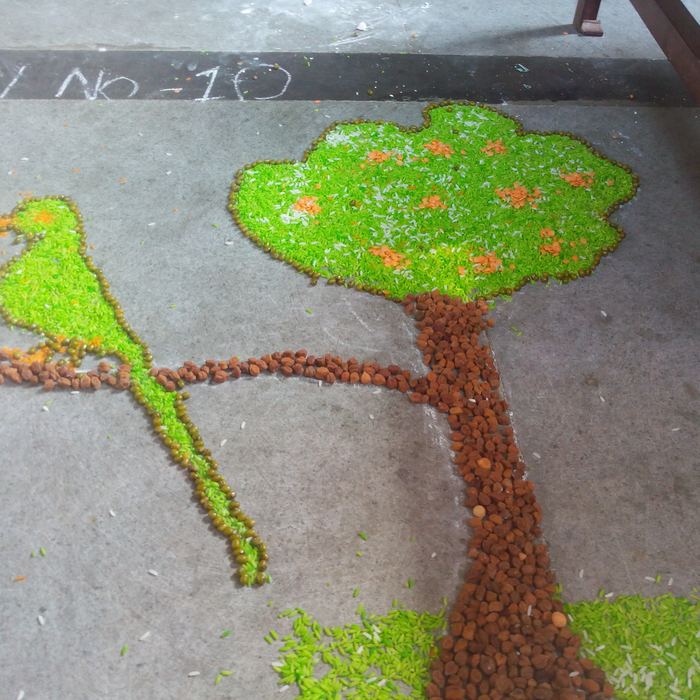 A grain rangoli of a green parrot perched on the branch of a broad tree, made from coloured rice, moong and chickpeas, with a roll number chalked beside it
