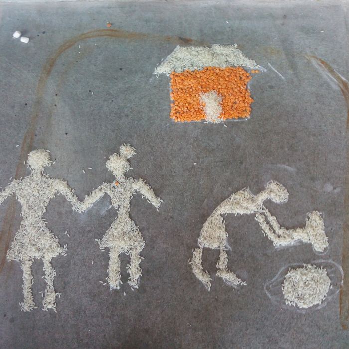 A grain rangoli showing figures of a family holding hands beside a house, outlined in plain rice with a roof and walls of orange lentils