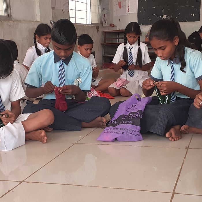 A long row of students in uniform sitting cross-legged on a tiled classroom floor, each working on a piece of knitting or crochet in green, red and white wool