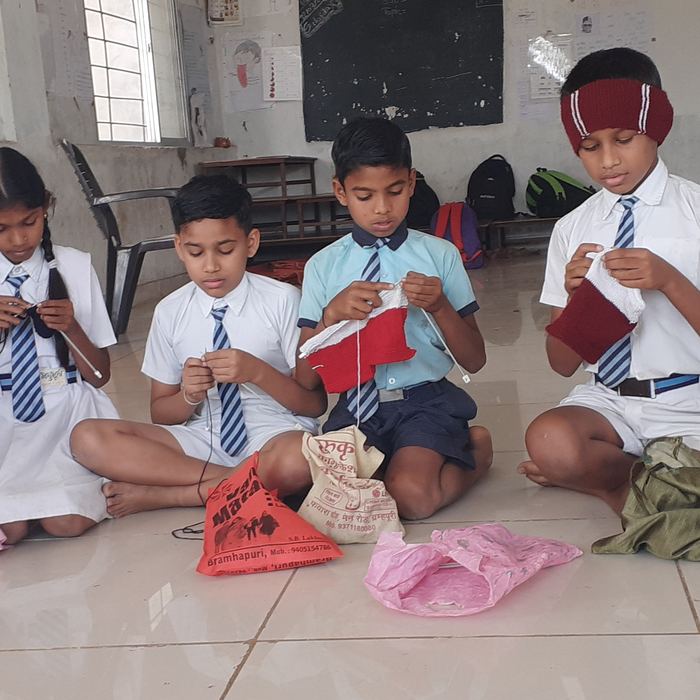 Seven students kneeling in a row on a tiled floor holding up part-finished knitted pieces, one boy wearing a maroon knitted headband