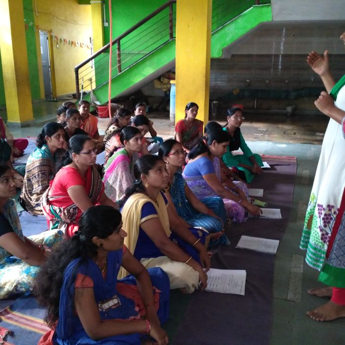 A large group of mothers in saris sitting on mats on the floor of the school hall, listening to a teacher speaking and holding printed handouts