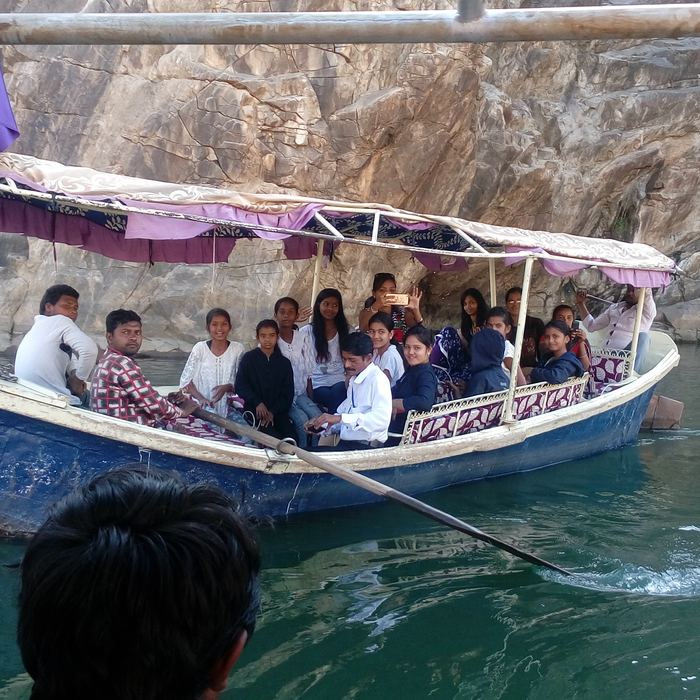 Students seated under a cloth canopy in a blue wooden boat being poled along green water beneath tall pale rock cliffs