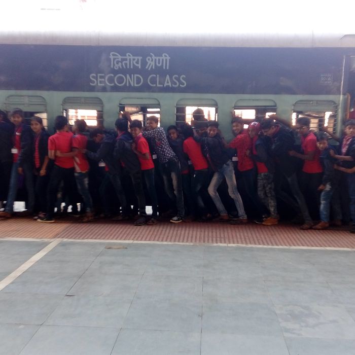 A long line of students in red and dark clothes with name badges boarding a green second class railway carriage from a station platform