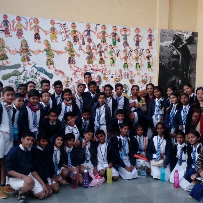 Students and teachers grouped in front of a museum wall covered with brightly painted tribal art figures of people, dancers and a peacock