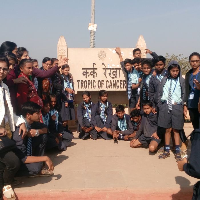 Students and teachers crowded around a stone marker reading Tropic of Cancer in Hindi and English, with green fields behind