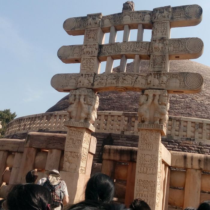 The heads of schoolchildren in the foreground looking up at an elaborately carved stone gateway with three architraves standing before the dome of a large ancient stupa