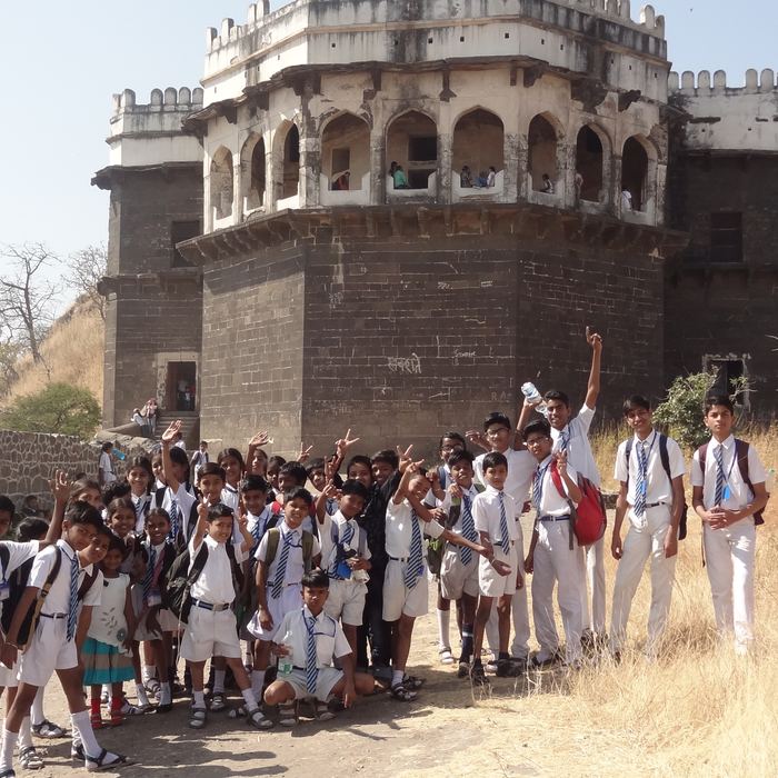 A crowd of students in white uniforms cheering with raised hands in front of a stone hill fort with arched upper galleries and battlements