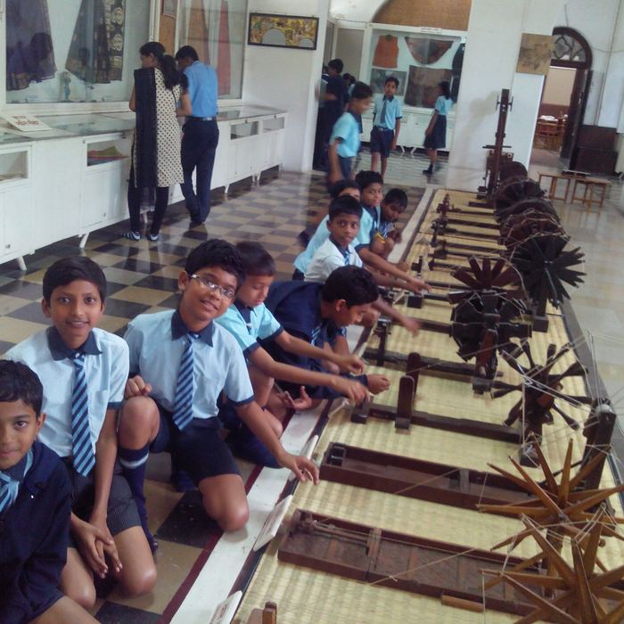 A line of students kneeling beside a long row of wooden charkhas and spinning wheels in a museum gallery, reaching out to turn them