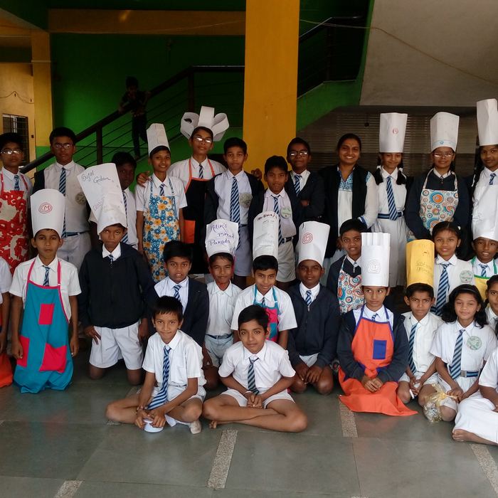 A large group of students wearing folded paper chef's hats and bright aprons posed together in the school hall with their teachers